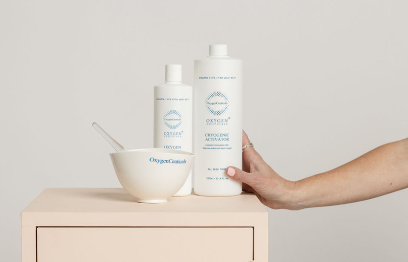 Hand reaches to hold a tall white OxygenCeuticals bottle next to a smaller bottle and a white bowl on a pale wooden surface.